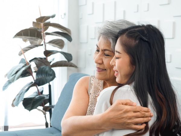 closeup portrait of happy asian senior mother or grandparent white hair embracing her beautiful daughter or grandchild with love care and comfort while sit on grey couch in living room at h