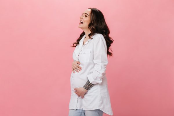 good humored pregnant woman in white shirt laughing on isolated cheerful girl in jeans smiling on pink background