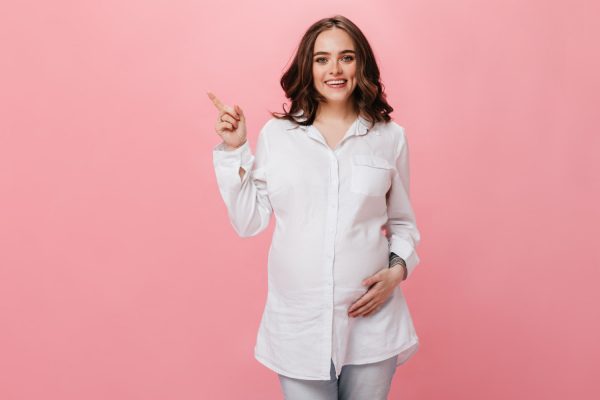 good humored pregnant woman touches belly and smile brunette girl in white shirt points to place for text on isolated pink background