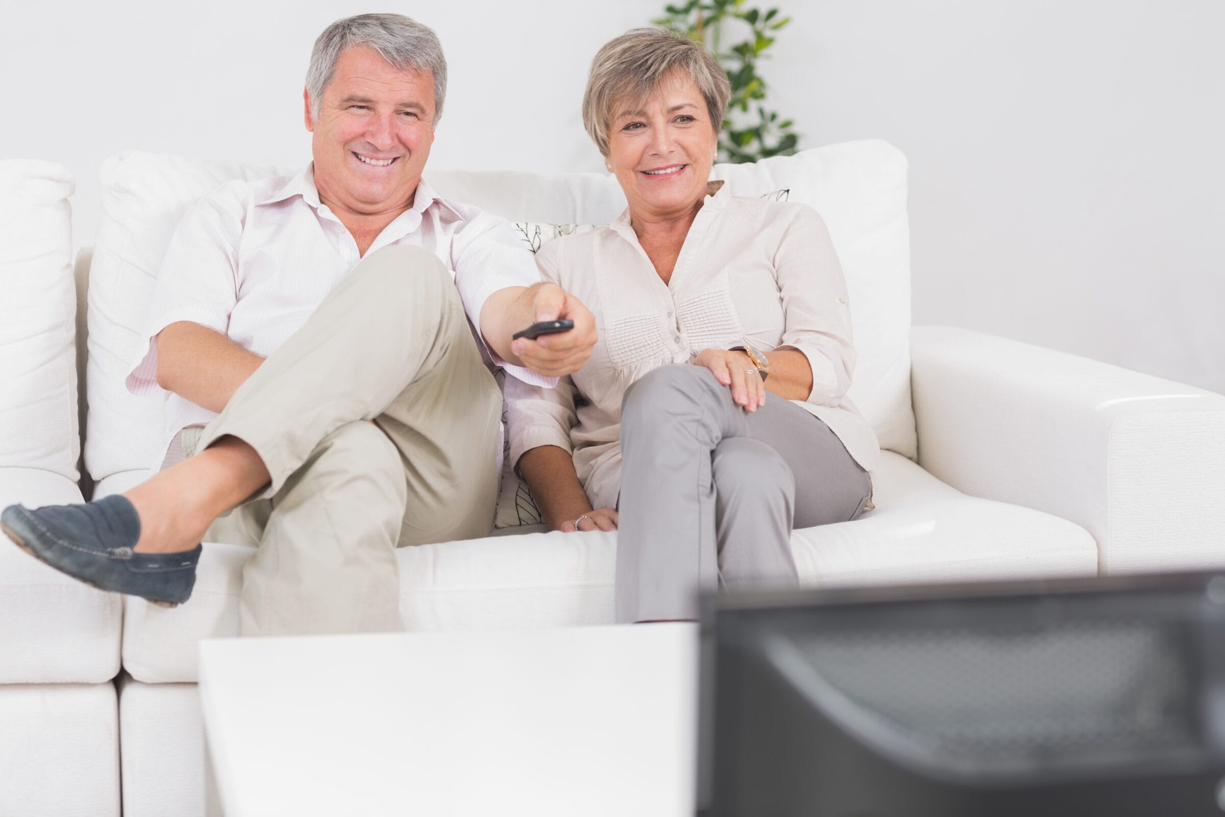 Casal de idosos assistindo TV em sofa brancoe controle na mão Desperta Débora