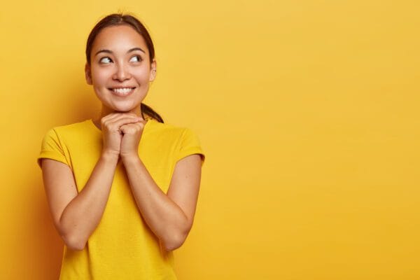portrait of dreamy satisfied ethnic girl concentrated aside with happy smile holds hands under chin looks with hopeful expression believes in better wears casual yellow t shirt stands in