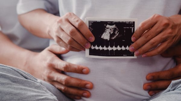 young asian pregnant couple show and looking ultrasound photo baby in belly mom and dad feeling happy smiling peaceful while take care child lying on sofa in living room at home